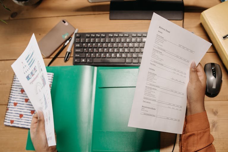 Hands holding and reviewing balance sheets over a desk with a keyboard and stationery.